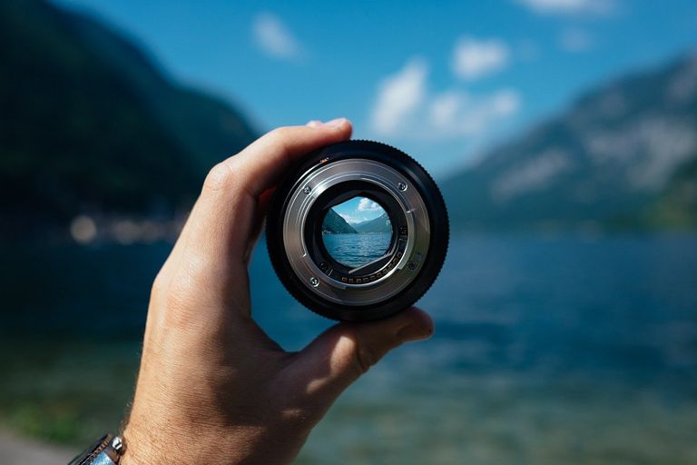 Eine Hand hält eine Objektivlinse durch die der Blick scharf auf das Panorama eines Bergsees gerichtet ist.