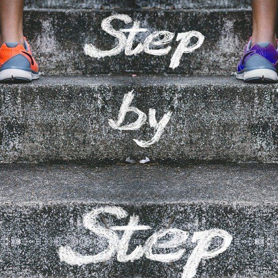Eine Steintreppe auf der zwei Jogger:innen nach oben laufen. Auf den Treppenstufen steht mit Kreide Step by Step geschrieben.