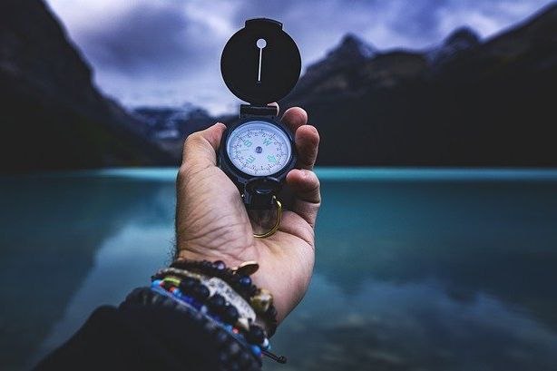 linke Hand mit einem Kompass vor dem Panorama  eines Bergsees und Bergen am Horizont