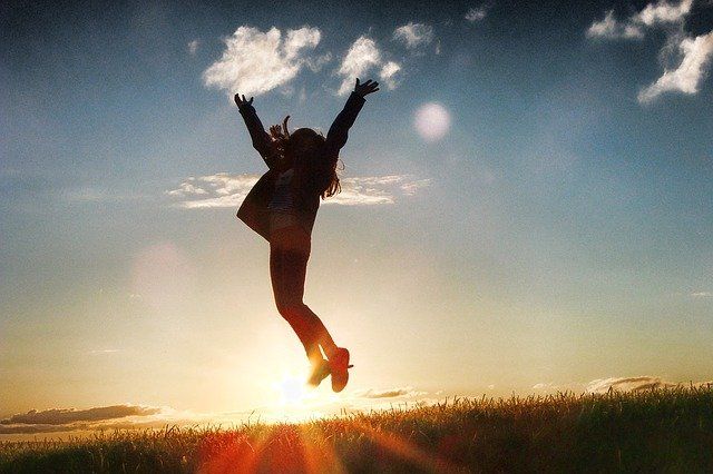 Eine Frau springt glücklich die Arme in die Luft werfend vor dem Panorama der untergehenden Sonne vor strahlend blauem Abendhimmel