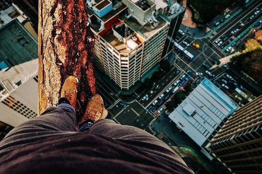 Ein Mensch balanciert auf einem Baumstamm zwischen zwei Wolkenkratzern mit der Perspektive über die eigenen Schuhe nach unten