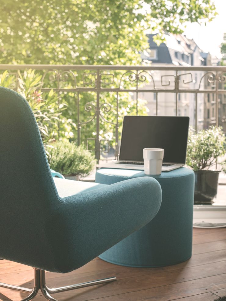 blauer Sessel mit Hocker, Laptop und Kaffeebecher auf einer Terrasse mit Blick ins Grüne